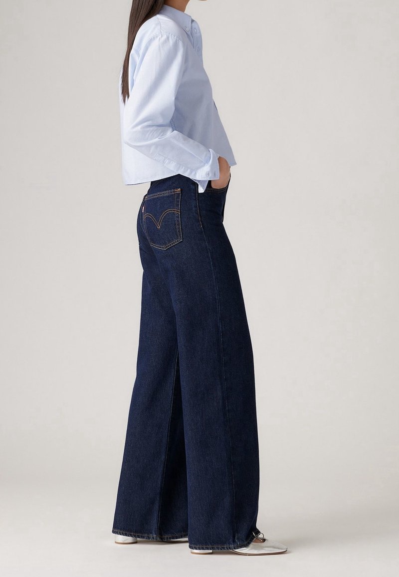 Chemise bleu clair à manches longues avec boutons, associée à un jean denim bleu foncé à jambes larges, avec des accents de surpiqûres sur les poches arrière. Chaussures argentées.