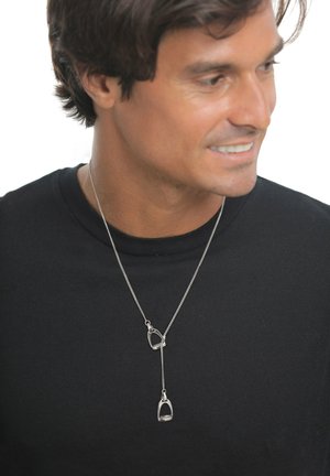 Mann lächelt und schaut nach rechts, trägt ein schwarzes Hemd und eine silberne Halskette mit zwei steigbügelähnlichen Anhängern.