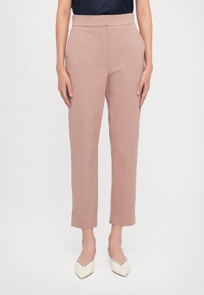 Mujer vistiendo pantalones de tiro alto y pierna recta en color malva y zapatos de punta blanca sobre un fondo blanco liso.