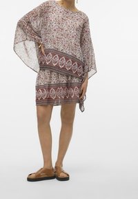 Robe transparente à motifs floraux en tissu léger, avec des manches chauve-souris et un bandeau décoratif contrastant à l'ourlet. Portée avec des sandales brunes.