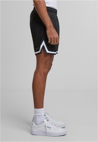 Shorts de sport noirs avec des rayures blanches sur les côtés et un ourlet en biais, fabriqués en tissu léger. Portés avec des baskets blanches et des chaussettes mi-mollet.