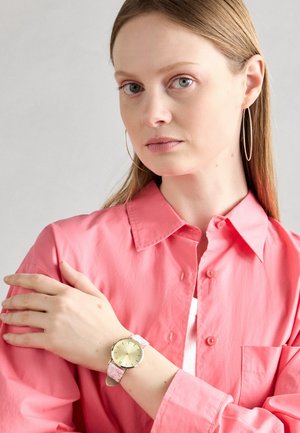 Kobieta o prostych jasnobrązowych włosach, nosząca duże kolczyki koła, różową koszulę na guziki oraz zegarek na nadgarstek z jasnoróżowym paskiem.