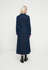 Lang denimkjole i mørkeblå med lange ærmer, krave og en figursyet talje. Har et flydende skørt og en glat tekstur. Sorte ankelstøvler.