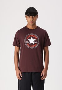 T-shirt en coton marron avec un grand logo circulaire blanc et orange Converse All Star sur la poitrine. Col rond, manches courtes.