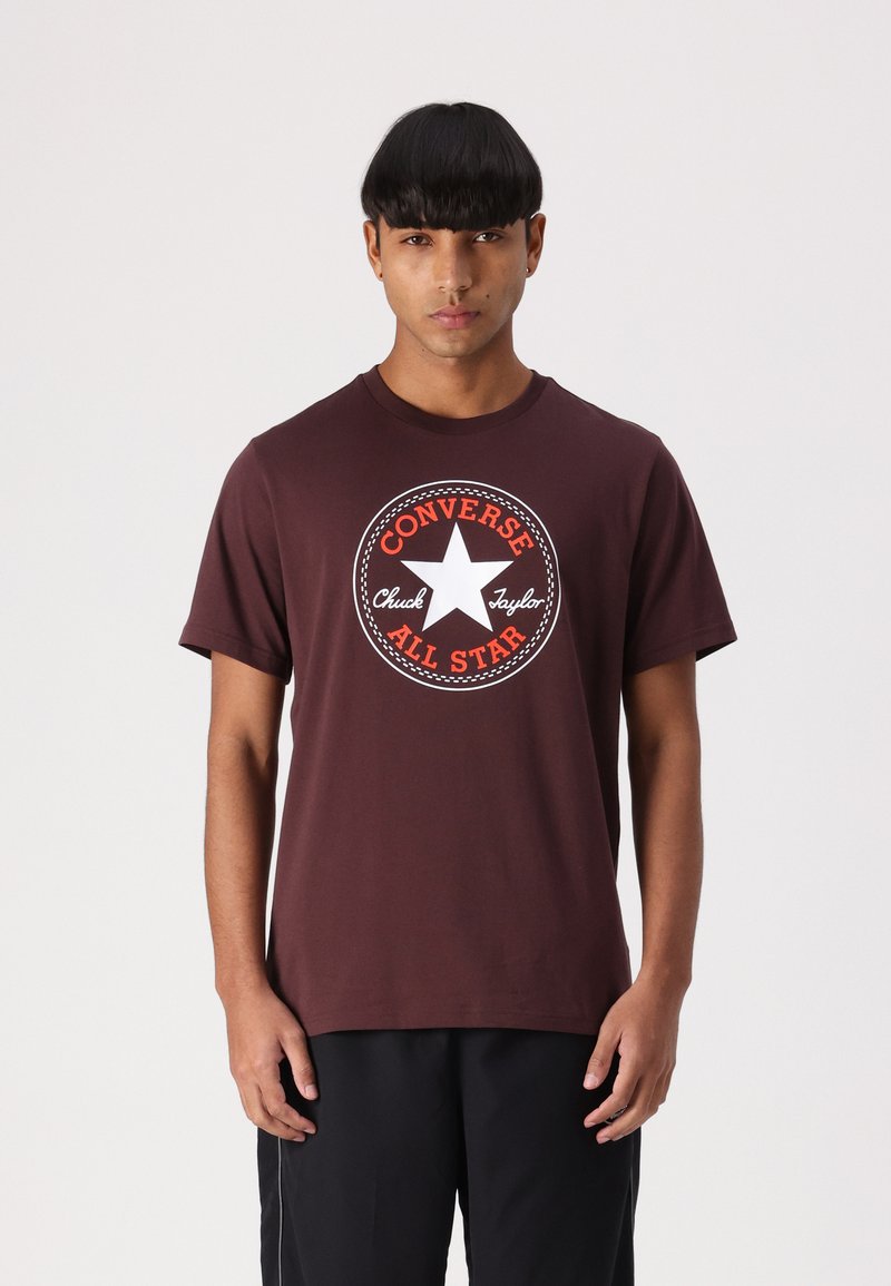 T-shirt en coton marron avec un grand logo circulaire blanc et orange Converse All Star sur la poitrine. Col rond, manches courtes.