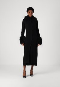 Zwarte geribbelde lange cardigan met veren kraag en manchet, voorzien van een knopenlijst en een getailleerd silhouet. Draagt over een gelaagde zwarte jurk.