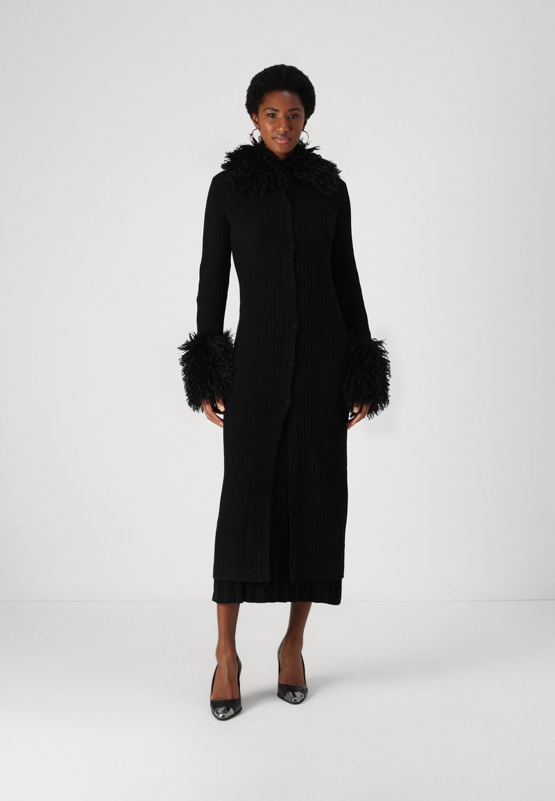 Zwarte geribbelde lange cardigan met veren kraag en manchet, voorzien van een knopenlijst en een getailleerd silhouet. Draagt over een gelaagde zwarte jurk.