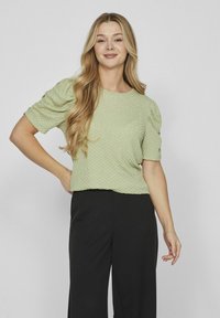 Lichtgroen textuurtop met korte puffmouwen, gecombineerd met nauwsluitende zwarte broeken. De stof vertoont een subtiel patroon door het hele oppervlak.