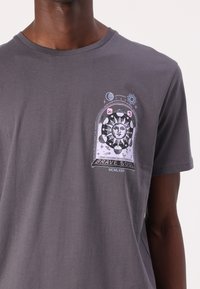 Grå bomulls-t-shirt med ett grafiskt tryck av en sol, planeter och texten "BRAVE SOUL" i rosa och svart på vänstra bröstområdet.