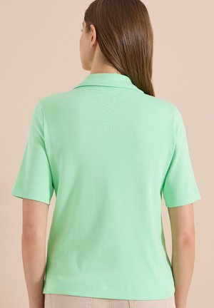 Kvinde med lige brunt hår iført en lysegrøn kortærmet poloshirt, vendt væk mod en beige baggrund.