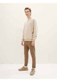 Mladý muž stojí, oblečený v béžové mikině, hnědých kalhotách, bílých ponožkách a béžových teniskách na jednobarevném světlém pozadí.