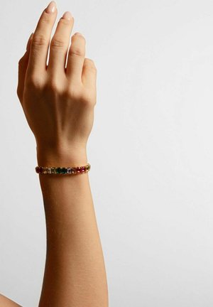 Armband in Goldoptik mit rechteckigen, bunten Edelsteinen in Grün, Rot und Klar, eingearbeitet in ein flexibles Band, getragen an einem Handgelenk.