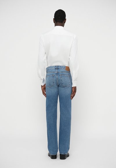 Bela srajca z dolgimi rokavi v kombinaciji z modrimi ravnimi jeans hlačami, z usnjeno nalepko na zadnjem delu. Gladka tekstura, sproščen kroj.