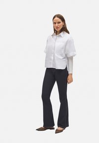 Mujer vistiendo una camisa blanca de manga corta superpuesta sobre una blusa de manga larga, pantalones oscuros acampanados y bailarinas puntiagudas de estampado de leopardo.