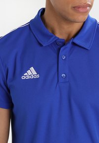 Blaues Poloshirt aus strukturiertem Stoff, mit einem kragen mit Knöpfen und drei Knöpfen. Weißes Adidas-Logo auf der linken Brust, mit weißen Streifen an den Ärmeln.