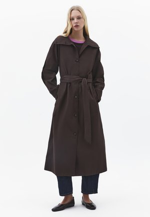 Frau steht mit den Händen in den Taschen und trägt einen langen dunkelbraunen Mantel mit Gürtel, dunkle Jeans, schwarze flache Schuhe und ein Oberteil mit violettem Kragen.