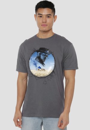 Man wearing gray t-shirt with graphic of a man in a hat and glasses above a desert scene with a van emitting blue smoke.