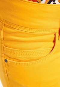 Los pantalones de algodón amarillos presentan un bolsillo frontal con costura decorativa, una textura suave y un pequeño remache metálico negro como acento.