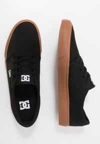 Black canvas sneakers with a brown rubber sole, featuring round laces and a logo on the tongue. Smooth texture, simple design.