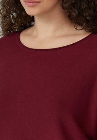 Burgunderfarbener Pullover mit gerippter Textur, weit geöffnetem Rundhals-Ausschnitt und weichem Material. Nahaufnahme mit Fokus auf den Ausschnitt und den Schulterbereich.