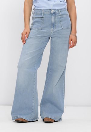 Vrouw in lichtblauwe wijde jeans en bruine schoenen, staand tegen een witte achtergrond met ontspannen handen langs haar zij.