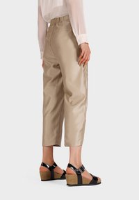 Pantaloni beige a vita alta con una texture liscia, abbinati a sandali con zeppa neri dotati di fasce in vernice e suola in sughero. Vista dal retro.