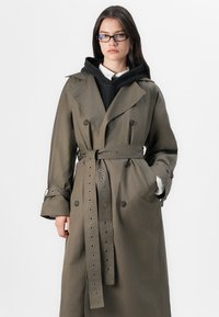 Olive roheline trench coat suure lapel'idega, topelt-nööbid ja vöödiga talje. Sisaldab silmadega detaile varrukatel ja taskutel.