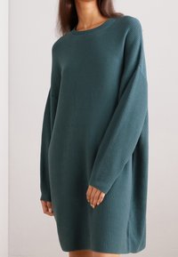 Abito lavorato a coste in teal con maniche lunghe, vestibilità comoda e spacchi laterali. La struttura è morbida e uniformemente distribuita, aumentando il suo calore.