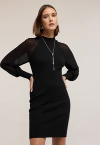 Schwarzes geripptes Kleid mit transparenten Puffärmeln und einem Mock-Neck. Besitzt eine taillierte Silhouette und eine silberne Kettenkette.