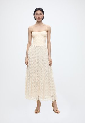 Γυναίκα με κοντά σκούρα μαλλιά που φοράει midi φόρεμα από εκρού δαντέλα χωρίς μανίκια και χρυσά ψηλοτάκουνα σανδάλια, στέκεται μπροστά σε ένα απλό λευκό φόντο.