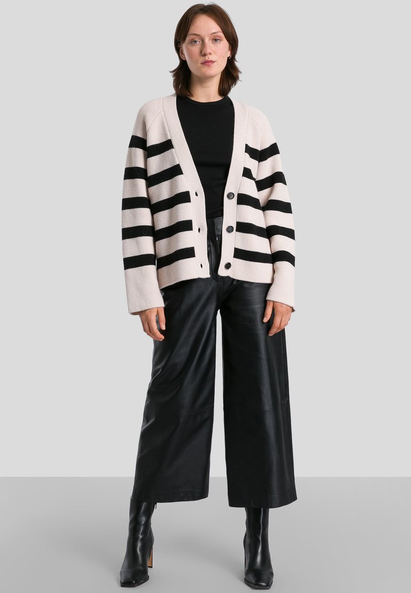 Cardigan a righe beige e nero, con un fit ampio, maniche larghe e bottoni sul davanti, abbinato a pantaloni in pelle neri a gamba larga.