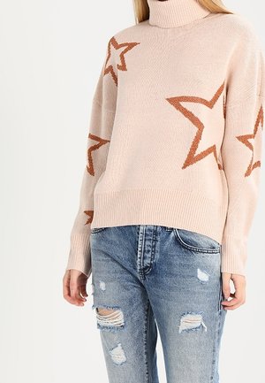 Maglione a maglia rosa chiaro con un collo alto e una vestibilità oversize. Presenta motivi a stella marroni, orlo a coste e maniche larghe. Indossato con jeans strappati.