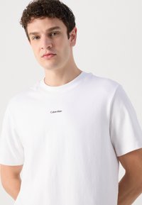 Biela bavlnená tričko s okrúhlym výstrihom a krátkymi rukávmi, s malým čiernym logom "Calvin Klein" potlačeným na hrudi.
