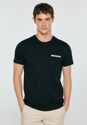 Noor mees, kes kannab musta T-särki väike trikolori triibuga rinnanohu kohal ja heledaid beeže pükse, seisab käed taskutes.