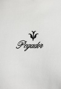 Țesătură albă cu text brodat negru "Pegdor" și un accent floral deasupra, evidențiind o textură netedă și un design simplu.