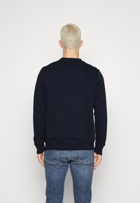 Persona con capelli biondi corti che indossa una felpa scura blu navy e jeans blu, in piedi di fronte a una parete bianca.