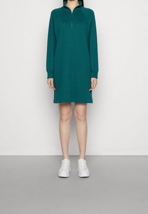 Žena v tyrkysově zelených šatech s dlouhým rukávem, zapínaných na zip u límce, a s bílými teniskami, stojí na pozadí s jednobarevnou bílou a šedou stěnou.