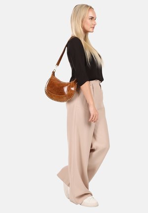 Femme aux longs cheveux blonds portant un haut noir et un pantalon beige à jambes larges, portant un sac à épaule en cuir marron, debout de profil.
