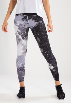 Femme portant des leggings noirs et gris à motif éclaboussé, des chaussures de sport noires et un haut sans manches blanc, se tenant devant un fond uni.