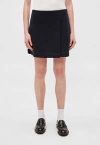 Zwarte mini-rok van gladde stof, met een subtiele ploo aan de voorkant, gecombineerd met zwarte loafers en witte sokken.