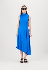 Asymmetrisches blaues Midikleid mit hohem Ausschnitt, ärmellosem Design, glatter Textur und fließendem Saum. Kombiniert mit schwarzen Sandalen.
