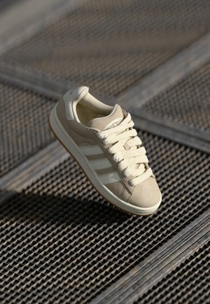 Beige kenkä valkoisilla raidoilla, kangas- ja mokkamateriaaleista valmistettu, jossa on teksturoitu pohja ja paksut nauhat, esiteltyä kuvioidun pinnan päällä.