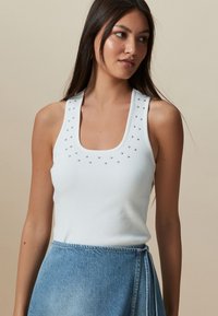 Débardeur blanc côtelé avec un décolleté carré et de petites clous argentés décoratifs le long des bretelles. Porté avec une jupe en jean.