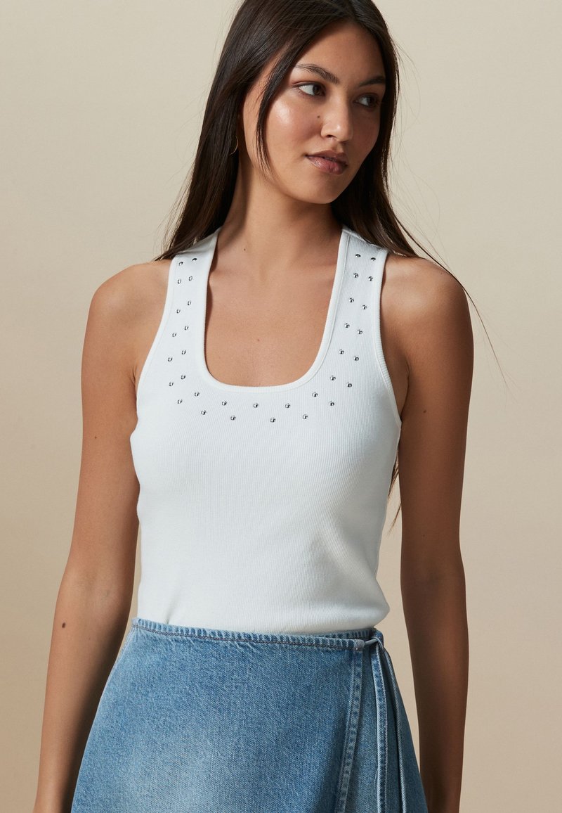 Débardeur blanc côtelé avec un décolleté carré et de petites clous argentés décoratifs le long des bretelles. Porté avec une jupe en jean.