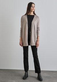 Cardigan beige clair en matière douce, avec une coupe décontractée et des poignets côtelés, associé à un pantalon noir slim et des bottines noires.