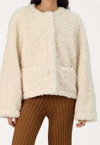 Krämfärgad, fluffig texturerad jacka med rund halsringning, långa ärmar och två framfickor. Matchad med ribbade bruna byxor.