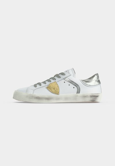 Λευκό sneaker χαμηλού τύπου με γκρι κορδόνια, χρυσό έμβλημα ασπίδας στο πλάι, μεταλλικό ασημί τακούνι και ελαφρώς φθαρμένη σόλα.