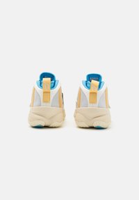 Atletische sneakers met een beige zool, een witte mesh bovenkant, gele en zwarte accenten, en een blauwe gewatteerde kraag. Stevige constructie met een trekhaak aan de achterzijde.