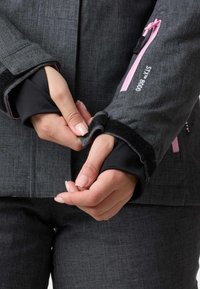 Grau textile Jacke mit einer Reißverschluss-Tasche mit pinken Akzenten und einer verstellbaren Velcro-Manschette, die auf einer Hand angezeigt wird, die die Manschette hält.