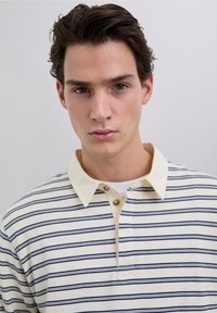 Polo con cuello en color crema con rayas horizontales en azul marino, cuenta con tres botones en el cuello y una tela suave y texturizada.